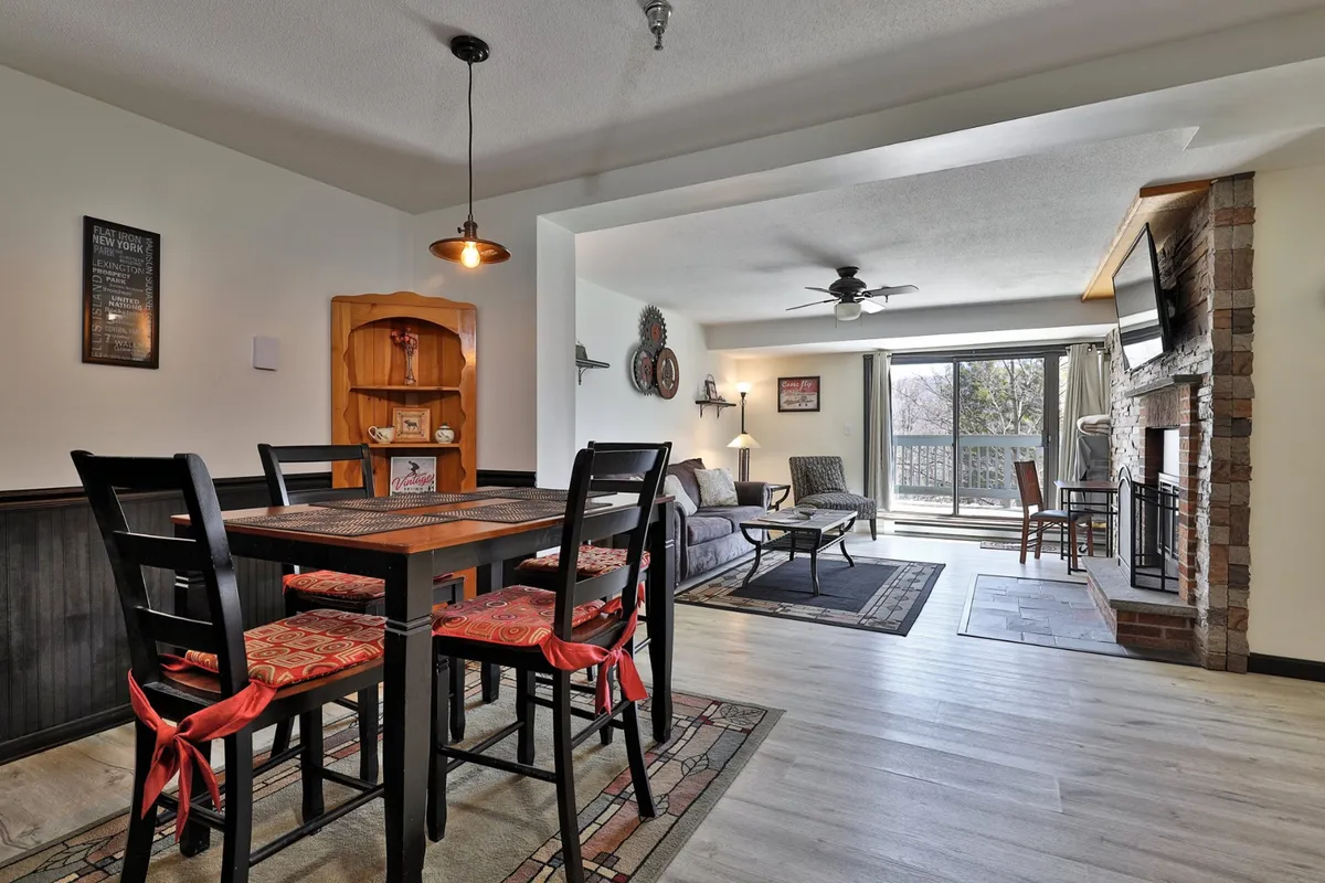 A spacious interior with a dining table, soft sofa, fireplace, and a large window letting in ample natural light. The setting is equally suited for winter fireside lounging or summer afternoons with fresh air and mountain views.