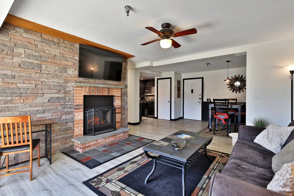 A cozy living room scene featuring a comfortable sofa, a flickering fireplace, a dining table set for gatherings, and a coffee table. The space is warmly lit, inviting, and perfect for relaxing or socializing. Large windows in the background hint at natural light and scenic views.
