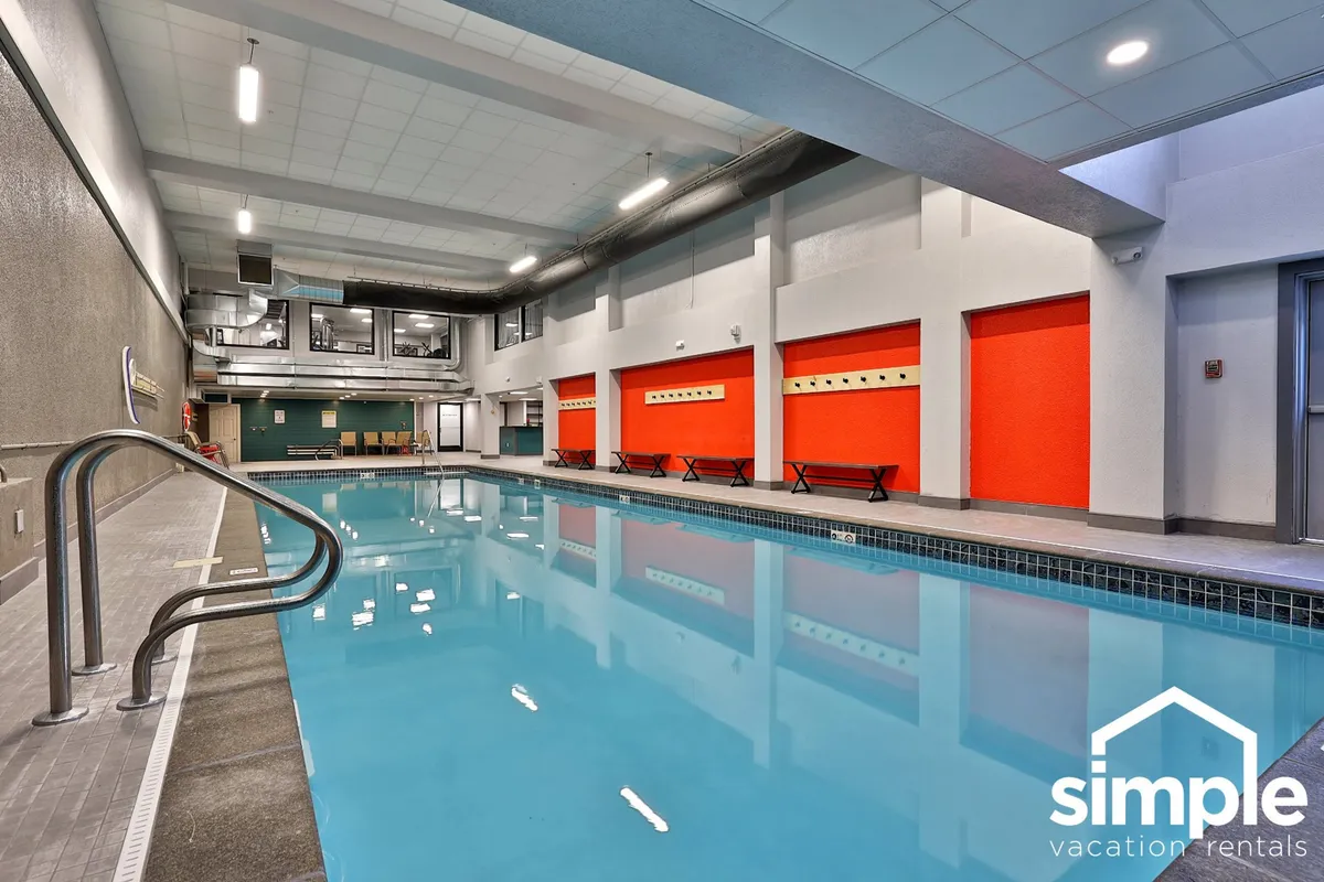 An inviting indoor pool scene featuring a swimming pool with red accent walls, a comfortable seating area, and soft lighting. The setting suggests year-round swimming and a relaxed space for both kids and adults to enjoy regardless of the weather.