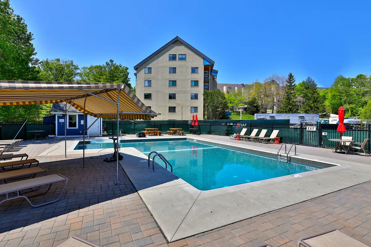 A sun-drenched outdoor swimming pool surrounded by lounge chairs, a canopy, and picnic tables. The scene includes several people relaxing on deckchairs and in the water, with a backdrop of lush greenery and clear skies, evoking the leisurely atmosphere of summer gatherings.
