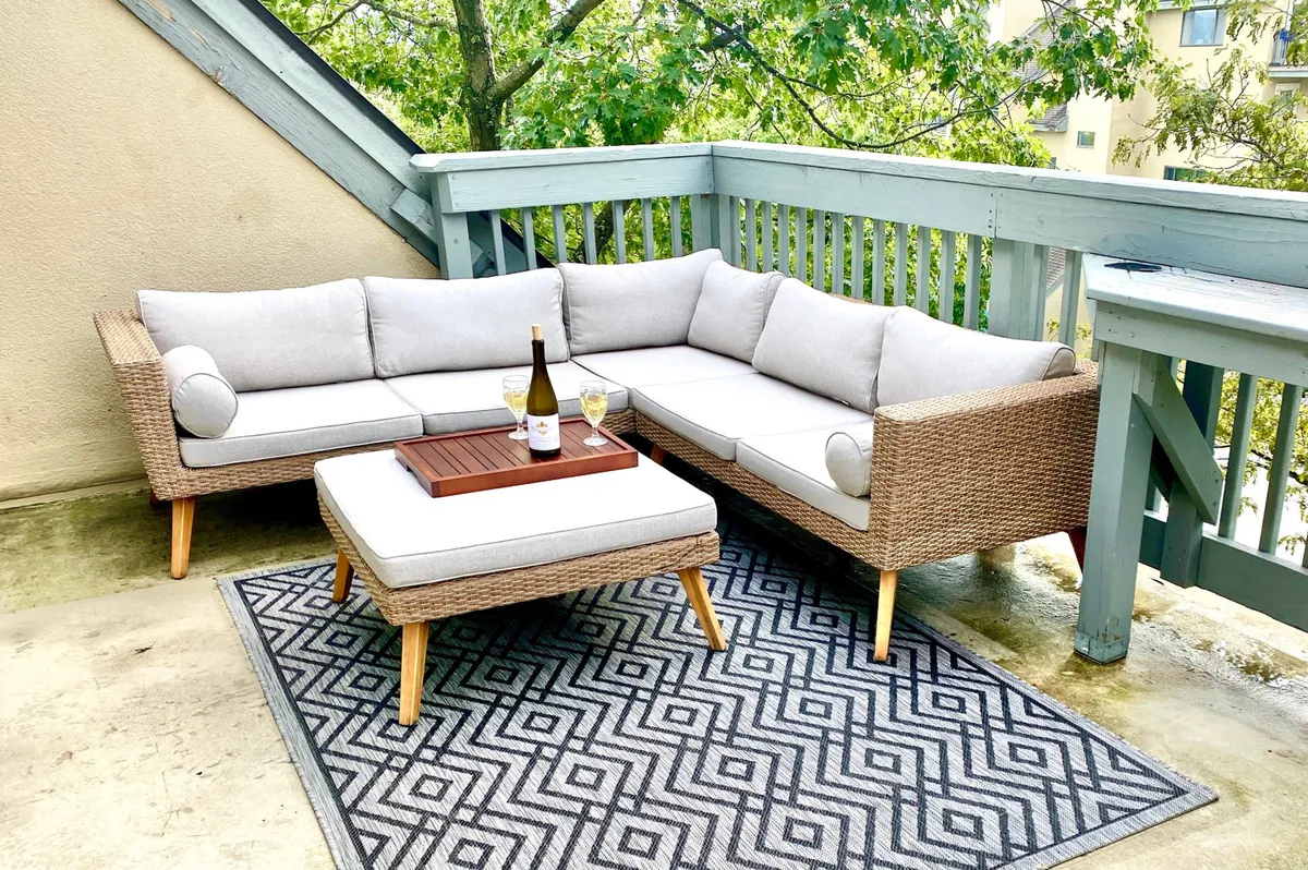 A comfortable outdoor seating area featuring a sectional sofa, coffee table with a wine bottle and glasses, and a cozy rug, all set on a patio with views of surrounding greenery. The arrangement suggests a relaxed, sociable environment ideal for unwinding or entertaining guests outdoors.