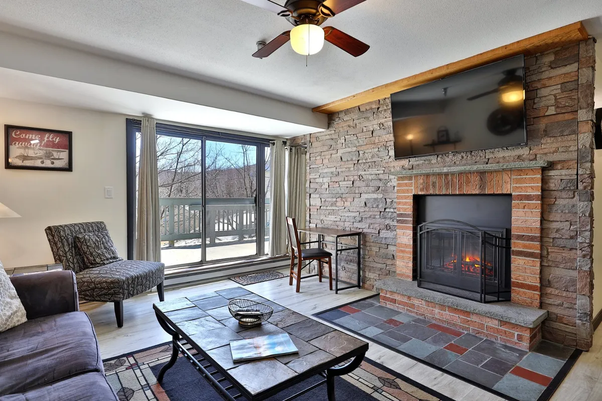 The image showcases a warm, inviting condo living room with a rustic stone fireplace as the centerpiece, flanked by comfortable seating and a cozy coffee table. Expansive windows allow natural light to fill the space, offering scenic views—likely of the snowy mountains—perfect for watching sunsets after a day on the slopes. The setting suggests a space ideal for relaxing, gathering with friends or family, or unwinding with a book by the fire.