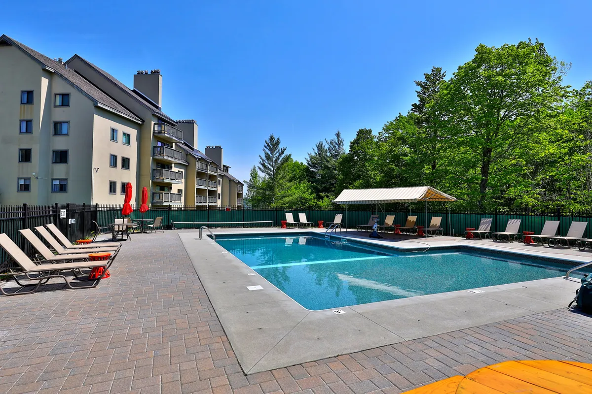 This exterior image captures the outdoor heated pool surrounded by lounge chairs, red umbrellas, a poolside cabana, and trees. Children are seen playing in and around the pool, creating a lively and inviting atmosphere ideal for family fun during the summer months.