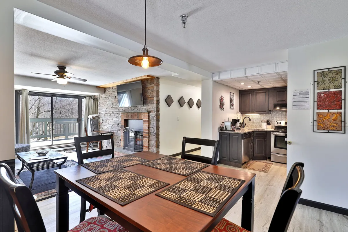 A cozy, open-plan dining and kitchen area with a dining table set for four, a nearby fireplace, and large windows bringing in natural light. The layout includes a view of the kitchen and invites gatherings and social meals.