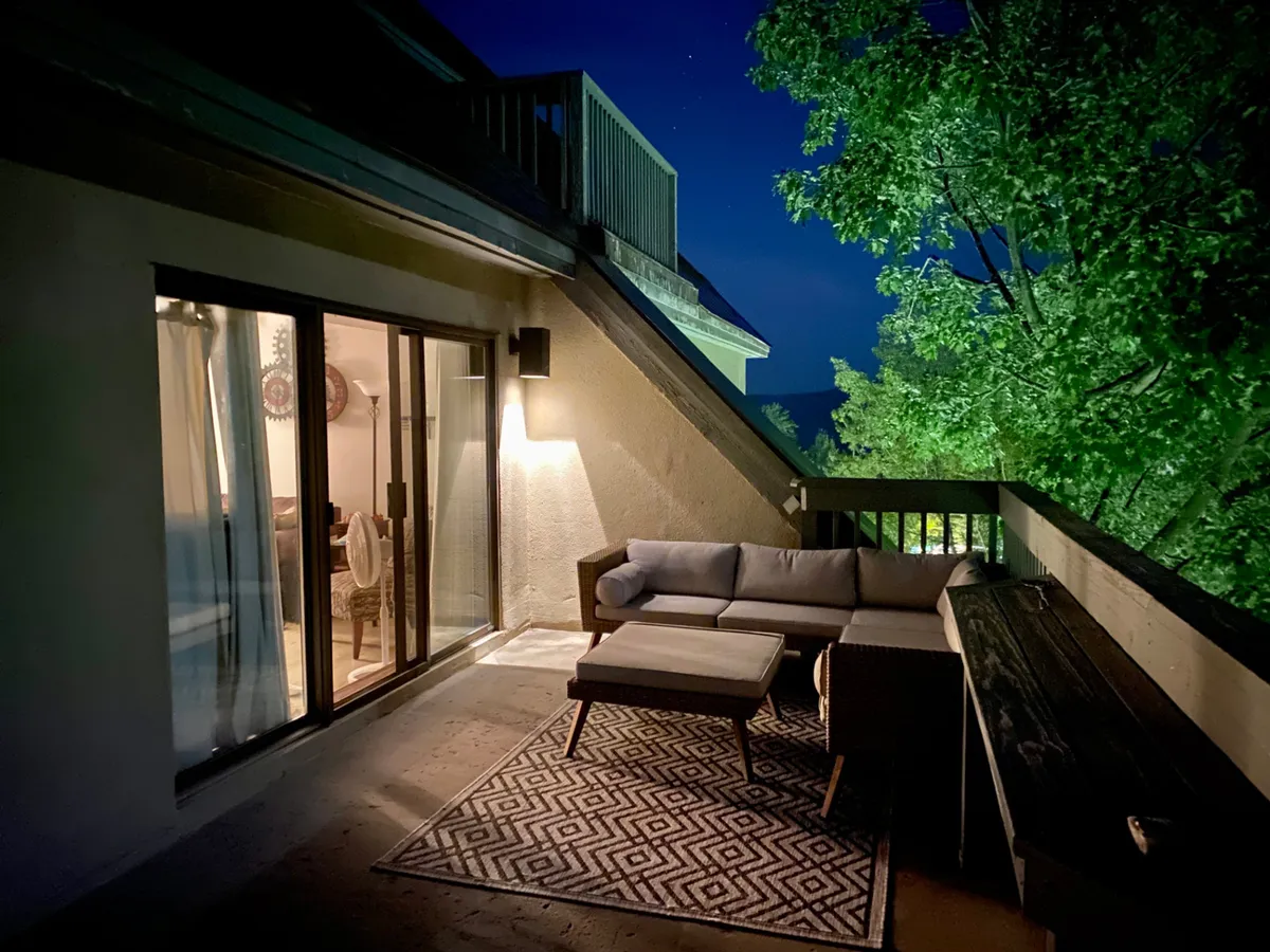 An inviting exterior scene featuring a cozy outdoor seating area surrounded by trees, with soft lighting spilling out from an illuminated interior space. The mood is calm, suggesting opportunities for evening relaxation or stargazing just steps from the door.